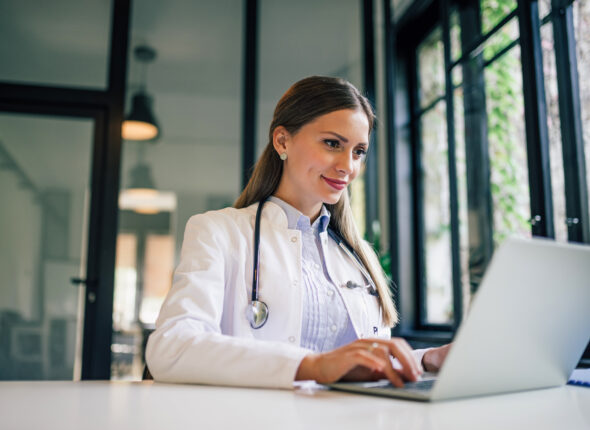 Positive doctor working on laptop in medical office, portrait.